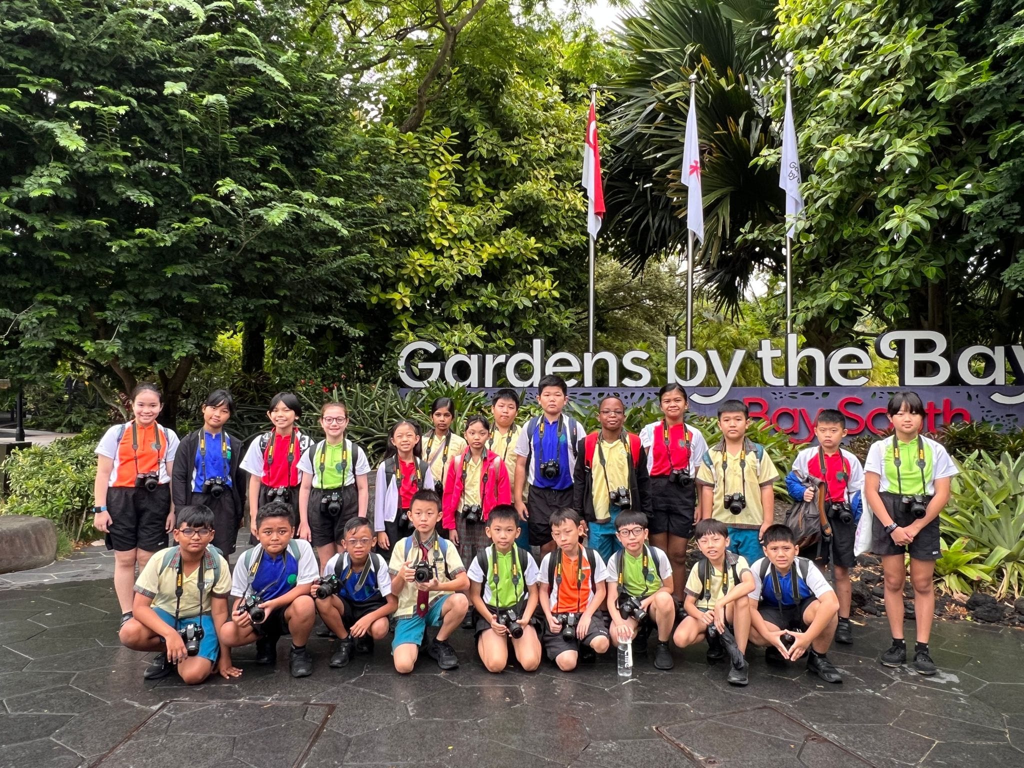A group of children with cameras pose in front of the Gardens by the Bay Bay South sign surrounded by trees.